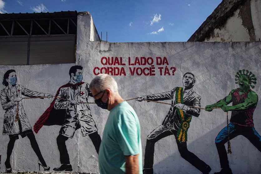 A man wears a face mask walks past by a mural depicting Brazilian President Jair Bolsonaro and a figure representing the coronavirus in a tug of war against health workers. Photo: Cris Faga/ZUMA Wire/dpa
